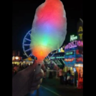 A glowing Flashing Fairy Floss Stick at a carnival, with colorful lights in the background.