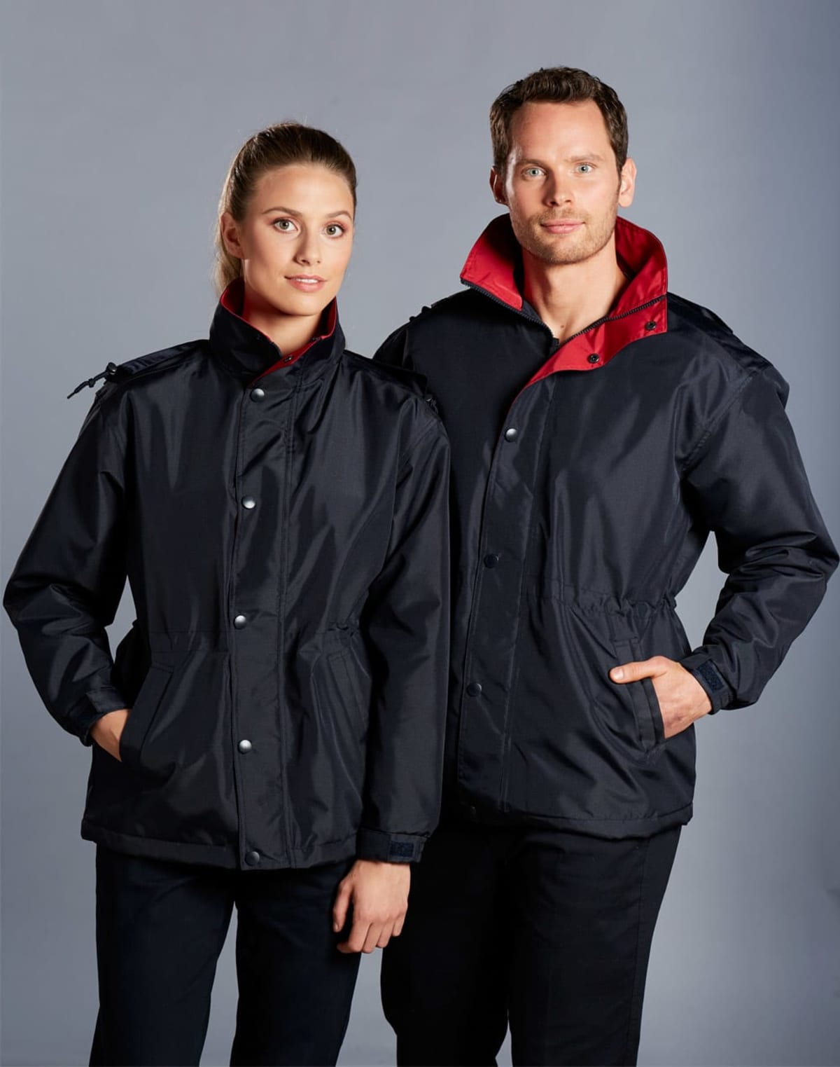 Two people wearing matching black jackets with red collars stand side by side, looking directly at the camera. The jackets have front pockets and are buttoned up. The background is a plain, neutral color.