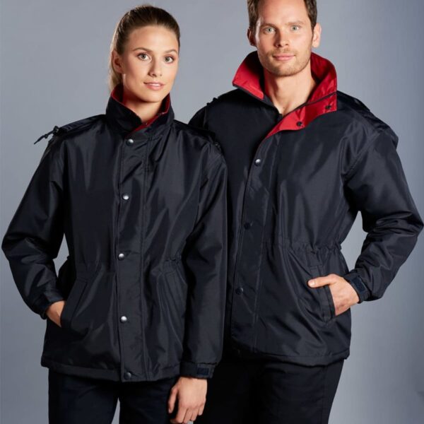 A man and a woman are standing side by side, both wearing matching black jackets with red collars. They have their hands in their pockets and are smiling slightly. The background is a plain gray color.