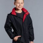 A smiling boy stands with his right hand in the pocket of his black jacket, which has a red collar. He is wearing a black shirt underneath and has short hair, posing against a plain gray background.