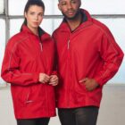 Two people standing side by side wearing matching red jackets with white piping. The person on the left has long dark hair tied back, while the person on the right has short hair and a beard. They are standing in front of a large window.