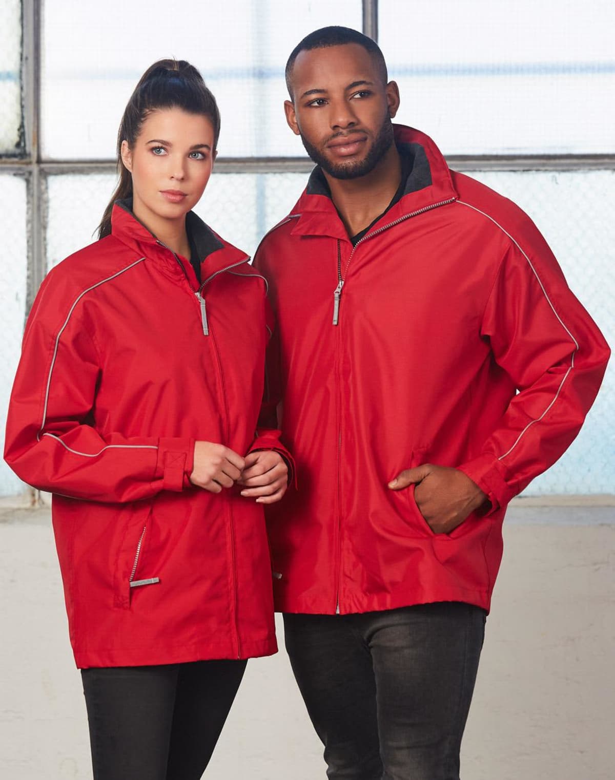 Two people standing side by side wearing matching red jackets with white piping. The person on the left has long dark hair tied back, while the person on the right has short hair and a beard. They are standing in front of a large window.