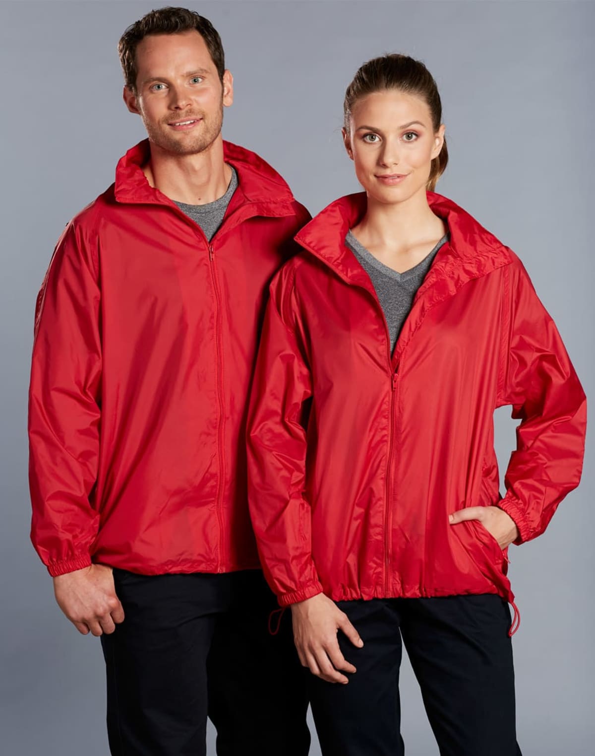 A man and woman wearing matching red jackets with gray shirts underneath pose against a neutral backdrop. The man has his arm around the womans shoulders. Both are smiling slightly and looking towards the camera.