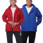 Two people standing against a white background. The person on the left is wearing a red jacket over a white shirt and black pants. The person on the right is wearing a blue jacket over a white shirt and black pants. Both are smiling.