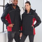 A man and a woman stand side by side wearing matching black and red jackets. The man has his hands in his pockets, while the woman has one hand on her hip. Theyre in a bright room by a large window.