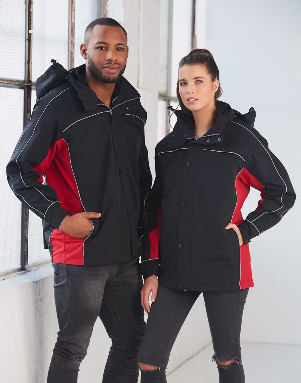 A man and a woman stand side by side wearing matching black and red jackets. The man has his hands in his pockets, while the woman has one hand on her hip. Theyre in a bright room by a large window.