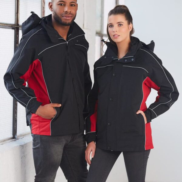 A man and a woman wearing matching black and red jackets stand indoors near a large window. Both have their hands in their pockets, facing the camera. The woman has her hair in a ponytail, and the man has a beard.