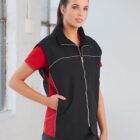 A woman with long hair in a ponytail is wearing a red shirt with a black zip-up vest. She is standing with one hand in her pocket and looking slightly to the side, against a light background.