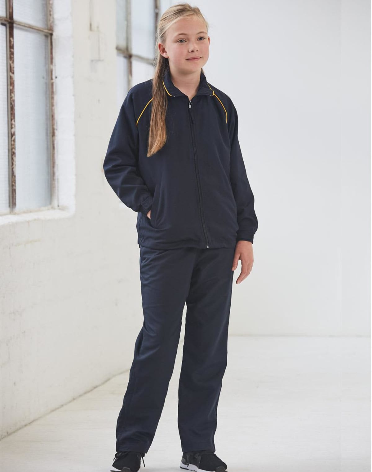A young person stands indoors wearing a navy tracksuit with yellow piping. They have long hair and are smiling slightly, with hands in their pockets. The room has white walls and a large window.