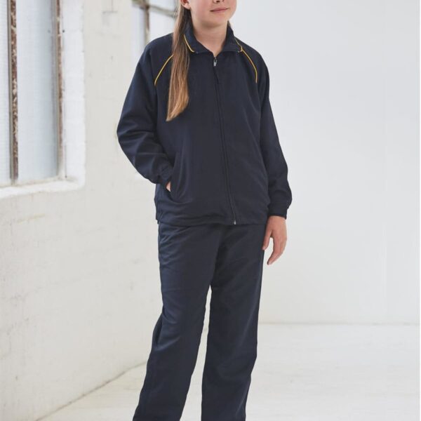 A person is standing indoors near large windows, wearing a dark tracksuit with yellow piping and black shoes. The setting is bright with natural light coming from the left.