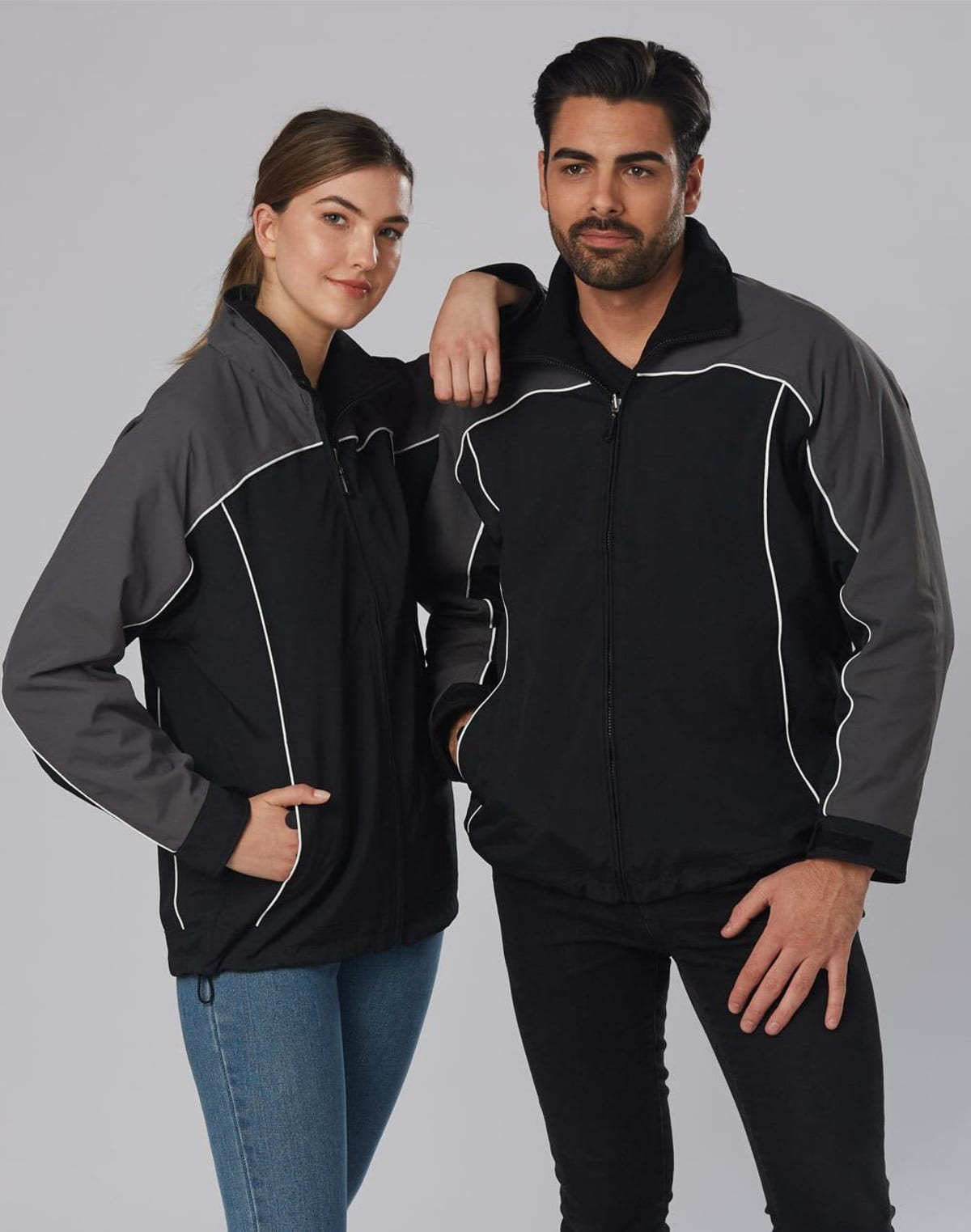 A woman and a man are posing together, both wearing matching black jackets with gray sleeves and white piping. The woman has one arm resting on the mans shoulder. She is wearing jeans, while the man is in black pants. They stand against a plain background.