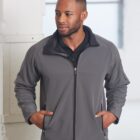 A person stands indoors wearing a gray zip-up jacket and a black shirt. They have short hair, a beard, and their hands in the jacket pockets. A large window in the background lets in natural light, creating a soft ambiance.