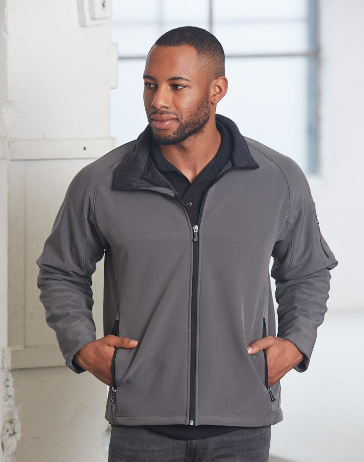 A person stands indoors wearing a gray zip-up jacket and a black shirt. They have short hair, a beard, and their hands in the jacket pockets. A large window in the background lets in natural light, creating a soft ambiance.