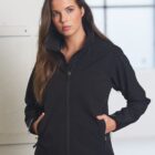 A person with long brown hair is wearing a black zip-up jacket with hands in the pockets. They are standing indoors against a light-colored wall and a window in the background.