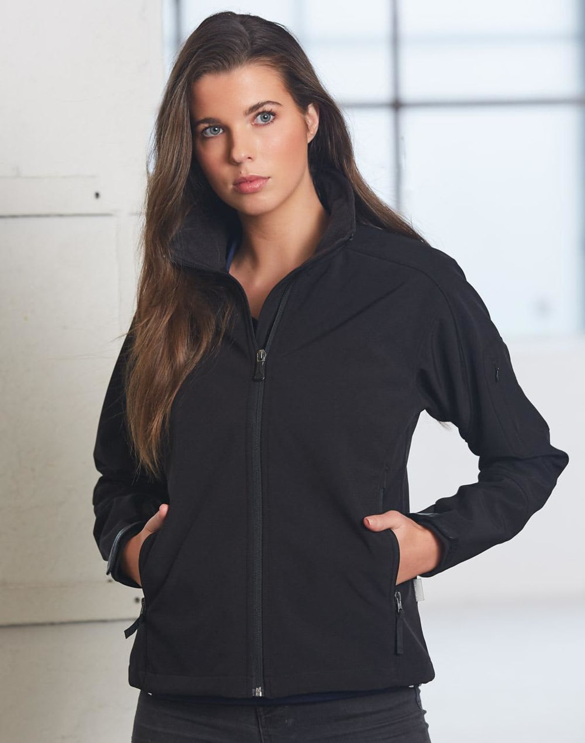 A person with long brown hair is wearing a black zip-up jacket with hands in the pockets. They are standing indoors against a light-colored wall and a window in the background.