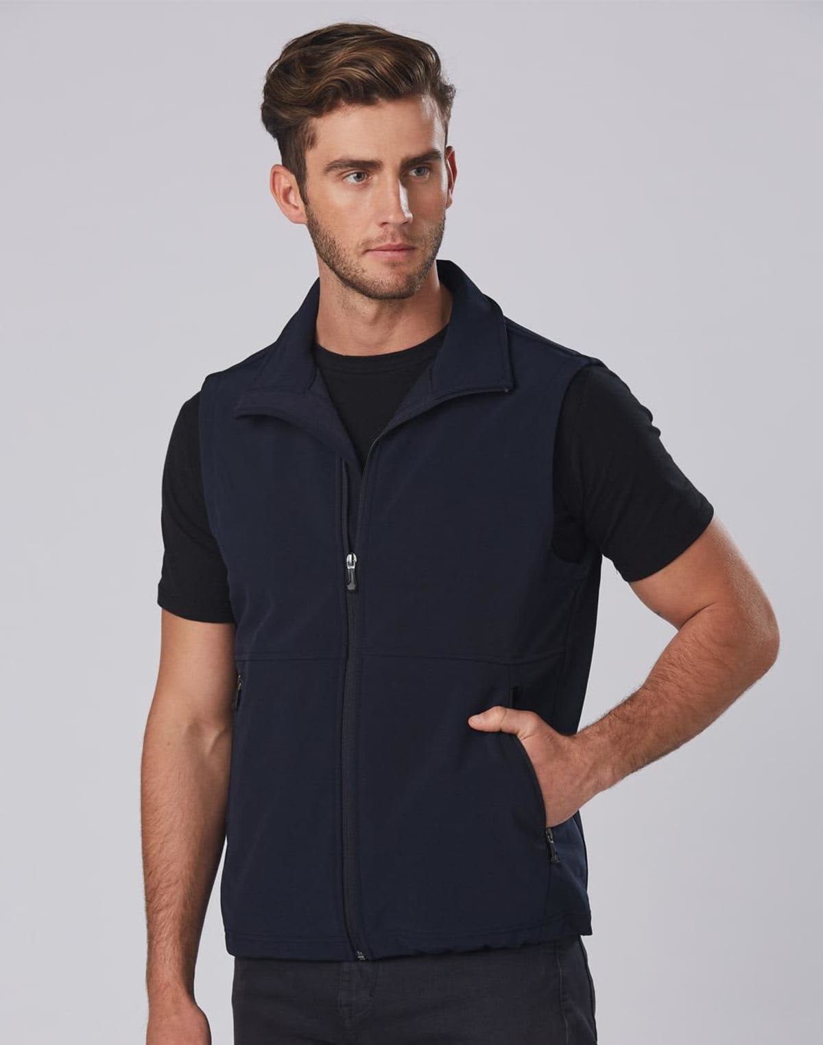 A man poses wearing a dark blue zip-up fleece vest over a black T-shirt. He stands with one hand in his pocket against a plain gray background.