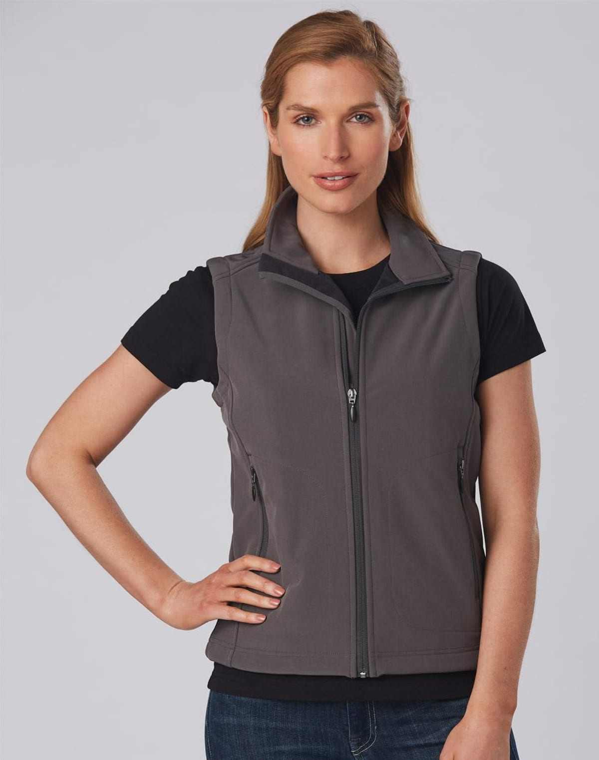 A person wearing a dark gray vest over a black T-shirt and blue jeans is standing against a plain background. They have one hand on their hip and are looking at the camera. The vest has a zippered front and zippered pockets.