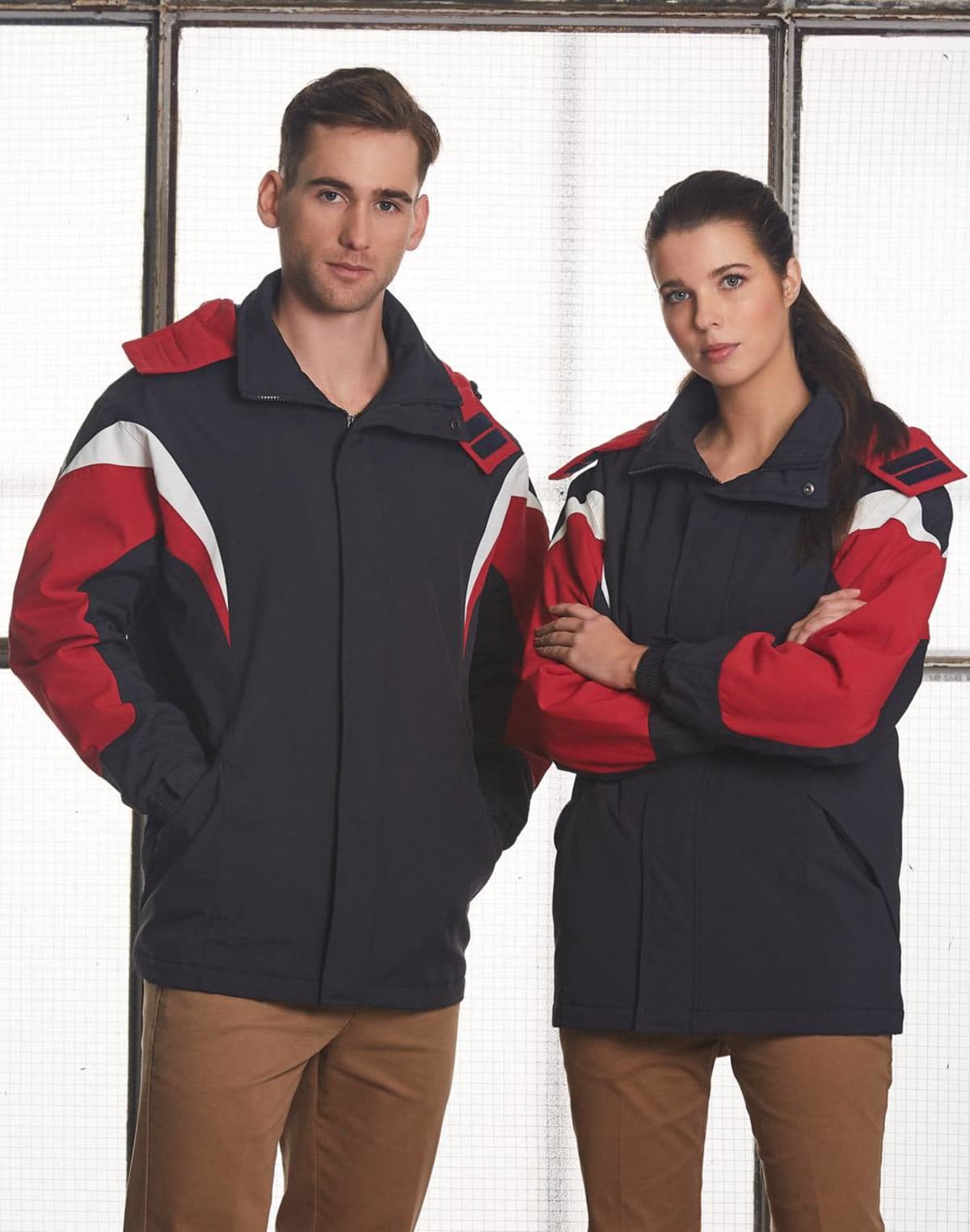A man and woman stand side by side, both wearing matching winter jackets with red, black, and white patterns. The man has his hands in his pockets, and the woman has her arms crossed. They are posed against a grid-like background.