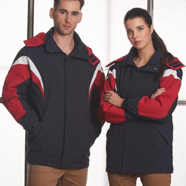 A man and a woman stand side by side, both wearing matching jackets with red, white, and black colors. They have neutral expressions and are in front of a window with a grid design. They are both wearing brown pants.