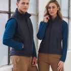 A man and woman stand side by side wearing dark vests over blue long-sleeve shirts and tan pants. They pose in front of large windows in a bright room. Both have their hands in their pockets, looking confidently at the camera.