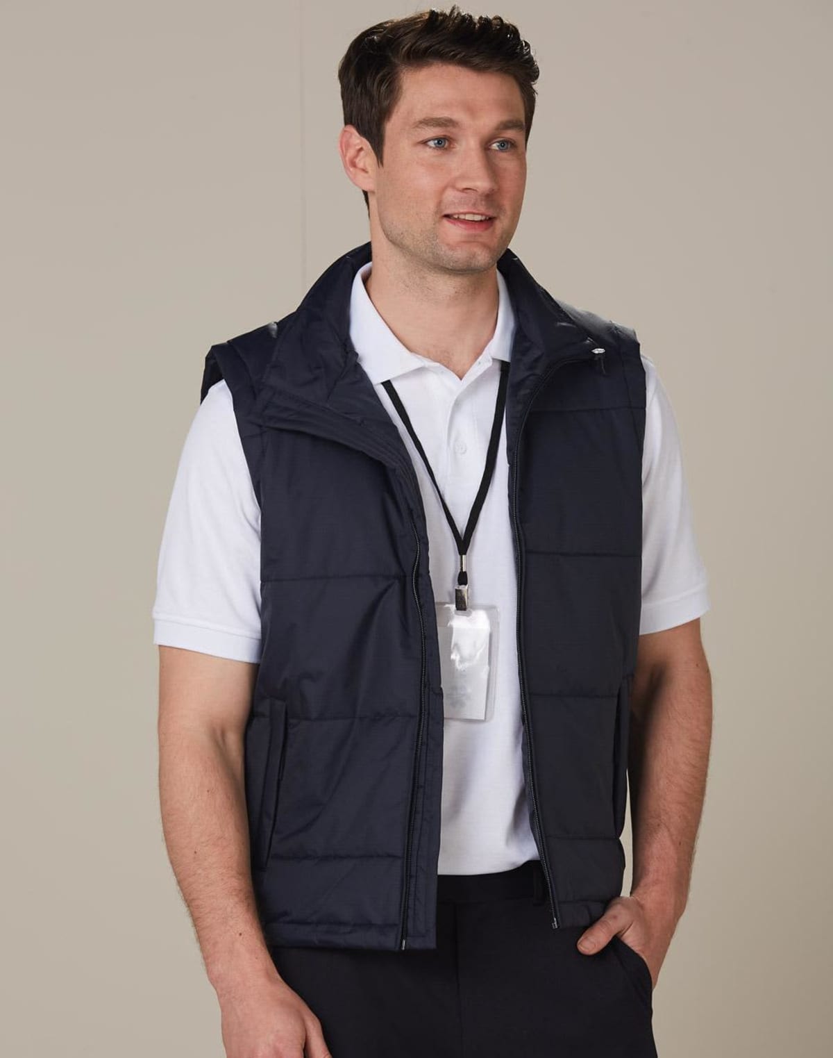 Man wearing a white polo shirt and black puffy vest with a lanyard and ID card around his neck, looking to the side and smiling. Neutral background.