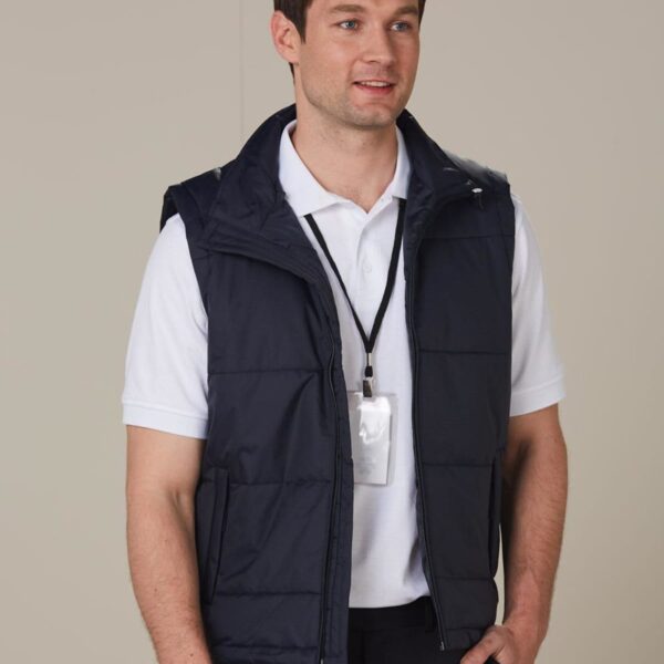 A man wearing a white polo shirt and a black padded vest stands against a plain background. He has a lanyard with an ID badge around his neck and is resting one hand in his pocket, smiling slightly.
