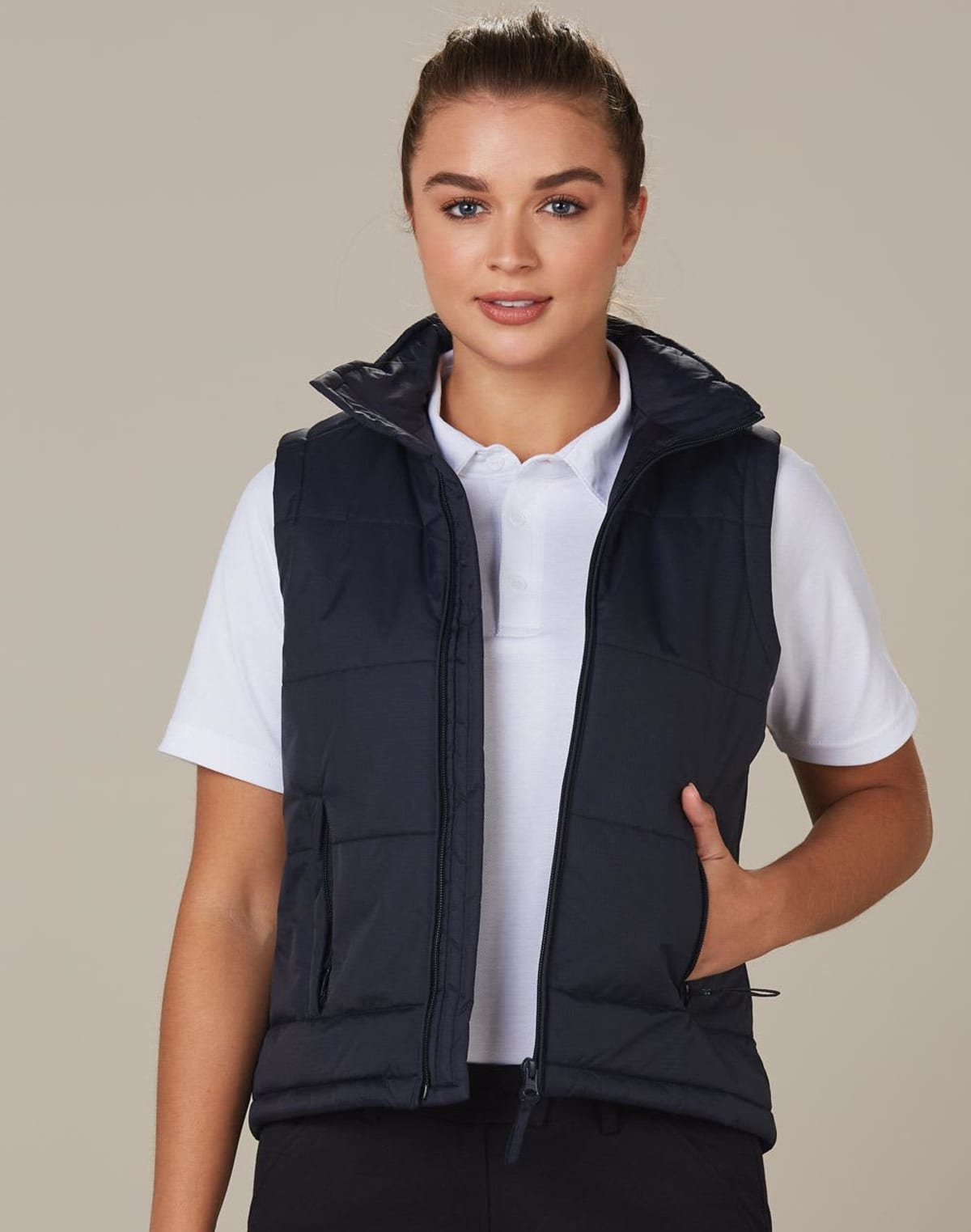 A person wearing a white polo shirt and a black puffer vest is posing against a neutral background. They have a hand in their pocket and are smiling subtly at the camera.