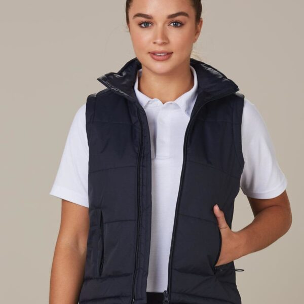 A person wearing a black puffer vest over a white polo shirt stands against a neutral background. The vest is sleeveless with a high collar, and they have one hand in a pocket, looking directly at the camera.