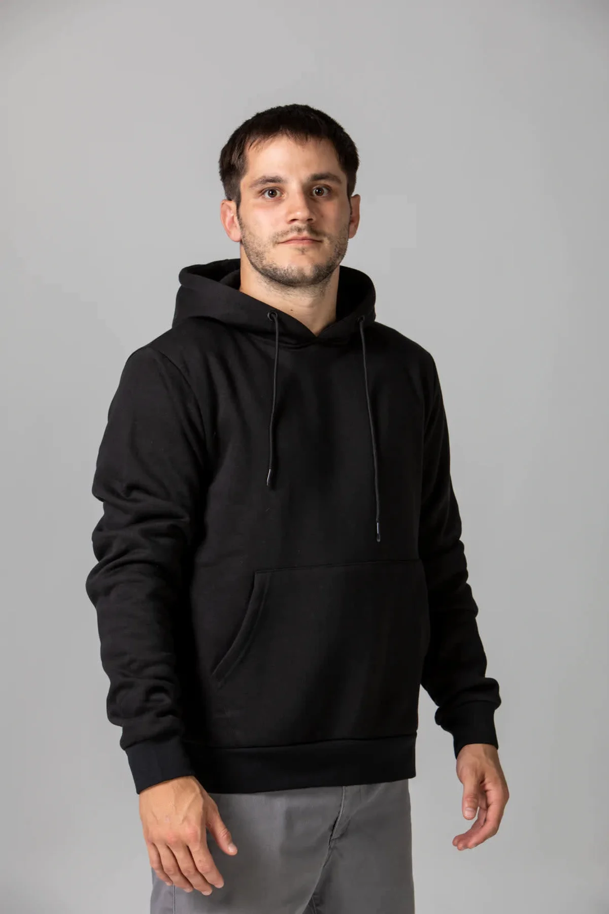 A person with short dark hair is wearing a black hoodie and gray pants, standing against a plain gray background. The individual is facing forward with a neutral expression.