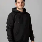 A man wearing a black hoodie and gray pants stands against a plain gray background. The hoodie has a front pocket and drawstrings. He is looking directly at the camera with a neutral expression.