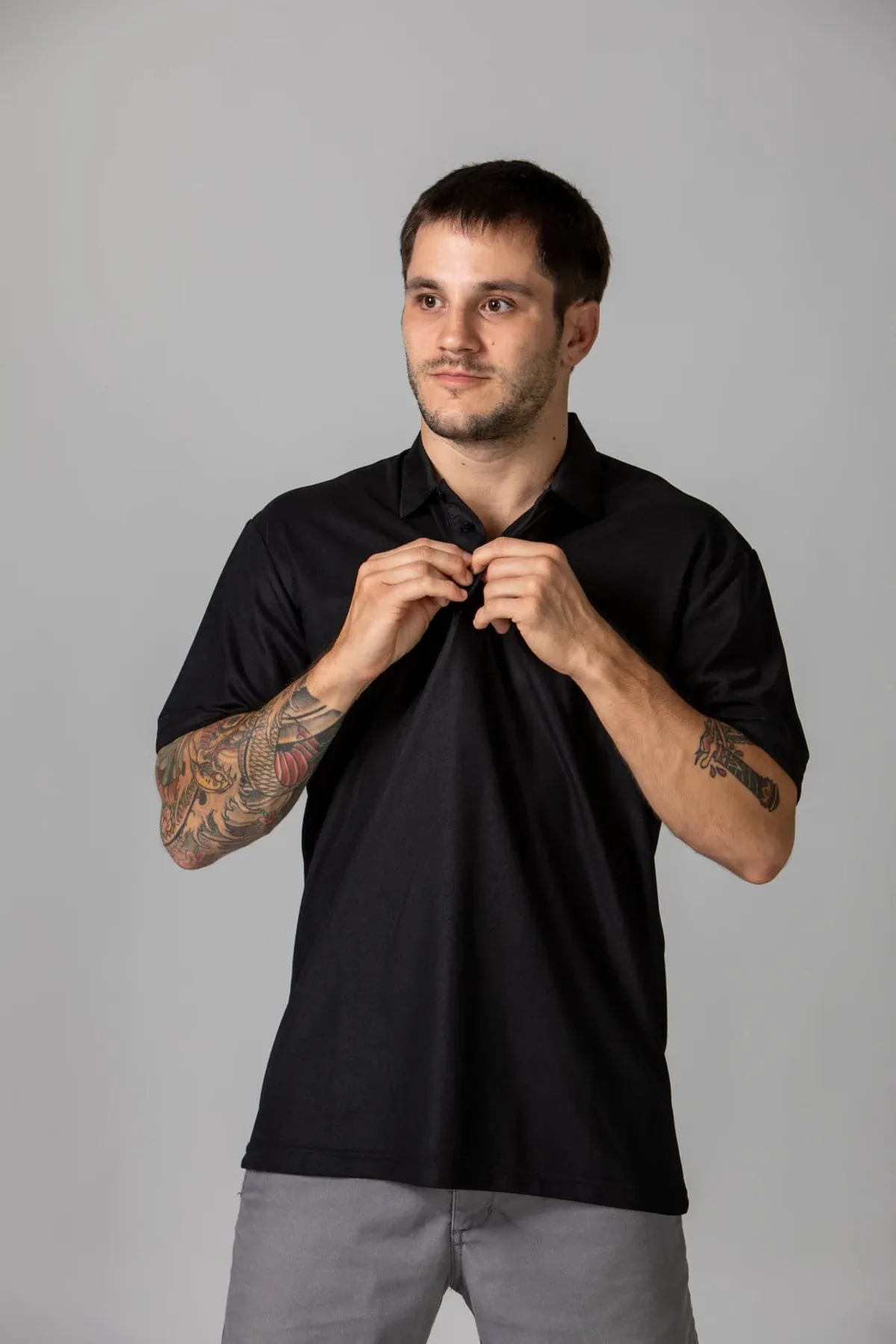 A man with tattoos on his arms is standing against a plain background, looking slightly to the side. He is wearing a black polo shirt and gray pants, adjusting the collar of his shirt with both hands.