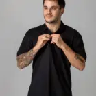 A person with tattoos on both arms is wearing a black polo shirt and gray pants. They are buttoning the shirt, and the background is a plain, light gray.