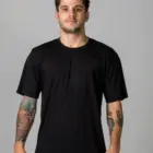 A man with short dark hair and tattoos on both arms wears a plain black T-shirt and gray pants. He stands facing forward against a plain gray background.