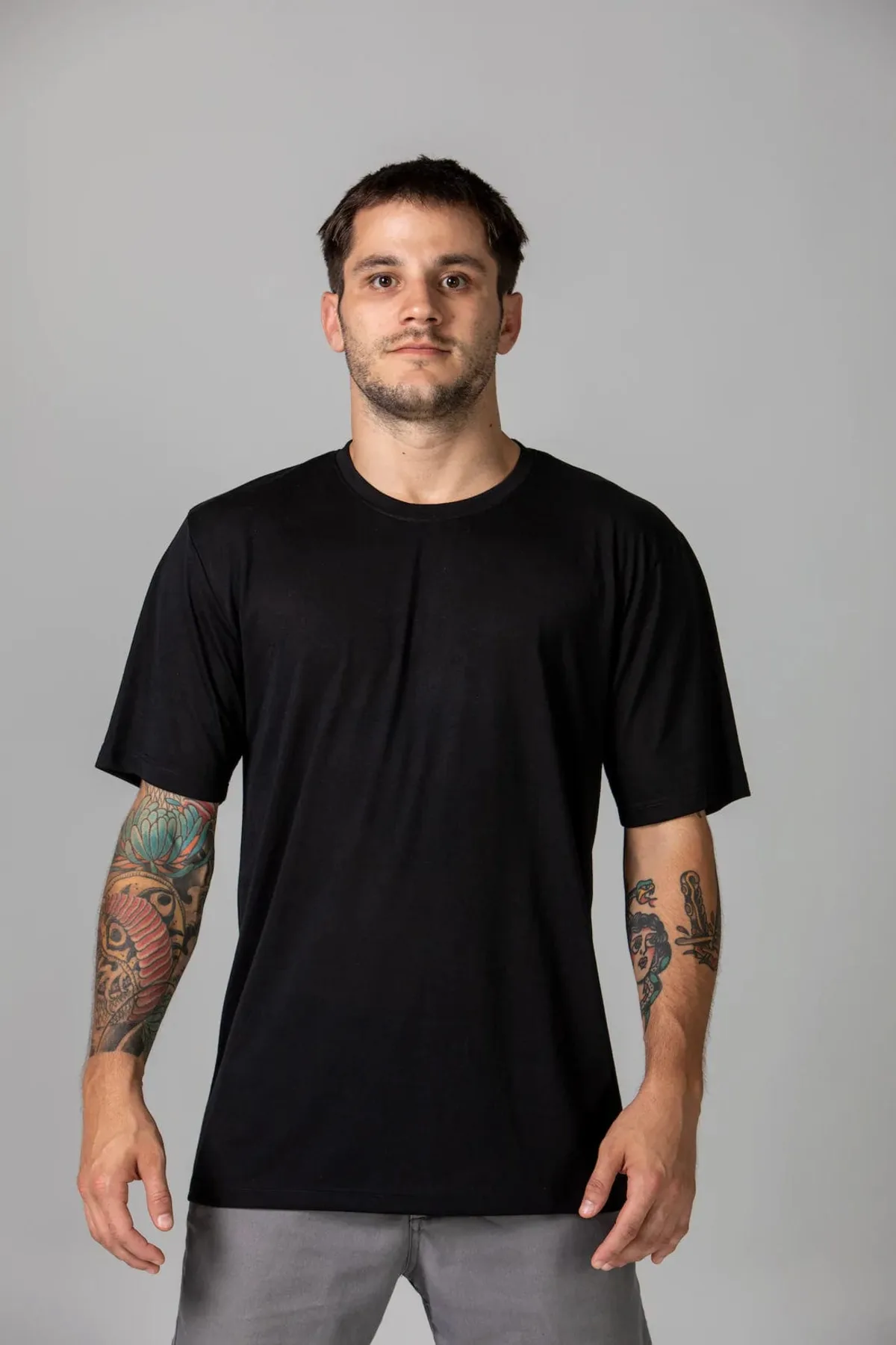 A man with short dark hair and tattoos on both arms wears a plain black T-shirt and gray pants. He stands facing forward against a plain gray background.