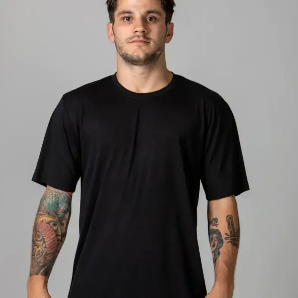 A man stands against a gray background, wearing a plain black T-shirt and gray pants. He has short hair and visible tattoos on his arms.