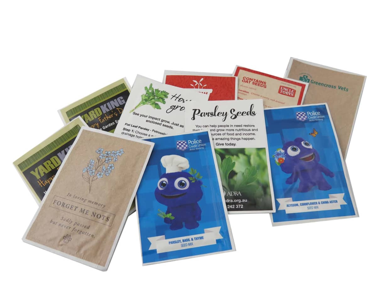 A collection of various seed packets, including designs with flowers, vegetables, and a cartoon character. Some packets feature text like Forget Me Nots and Pawsley Seeds. The packets are arranged casually on a white background.