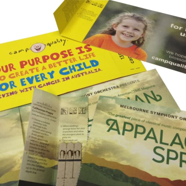 Various brochures are spread out. One from Camp Quality, highlighting its purpose to support children with cancer in Australia, with a smiling child on it. Another promotes the Melbourne Symphony Orchestras Appalachian Spring performance.