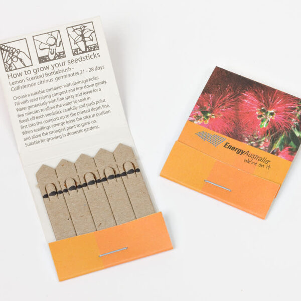 Open matchbook-style pack with five plantable seed sticks inside. The cover shows red flowers and has Energy Australia branding. Instructions for growing Lemon Scented Bottlebrush seeds are printed inside.