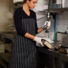 A person wearing a striped apron and gloves washes a pan using a sprayer in an industrial kitchen. The person is focused on the task, surrounded by stainless steel sinks and tiled walls.