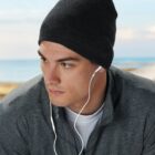 Man wearing a beanie and earphones, gazing to the side by the seaside.