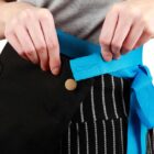 Person tying a blue belt around a black apron with striped pants in the background.