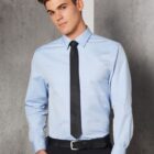 A man wearing a light blue dress shirt with a black tie and black pants stands against a gray wall, looking slightly to the side with a neutral expression.