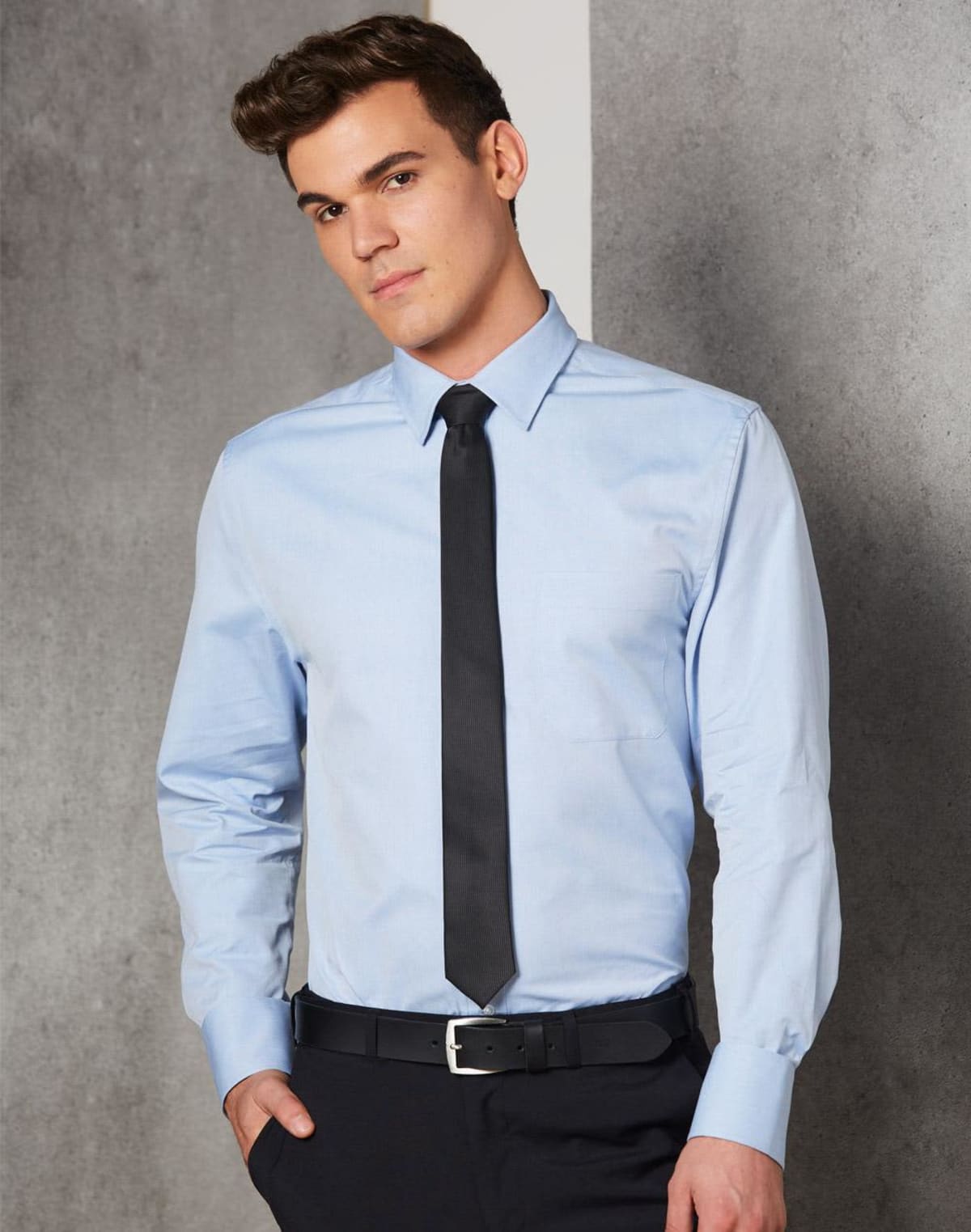 A man wearing a light blue dress shirt with a black tie and black pants stands against a gray wall, looking slightly to the side with a neutral expression.
