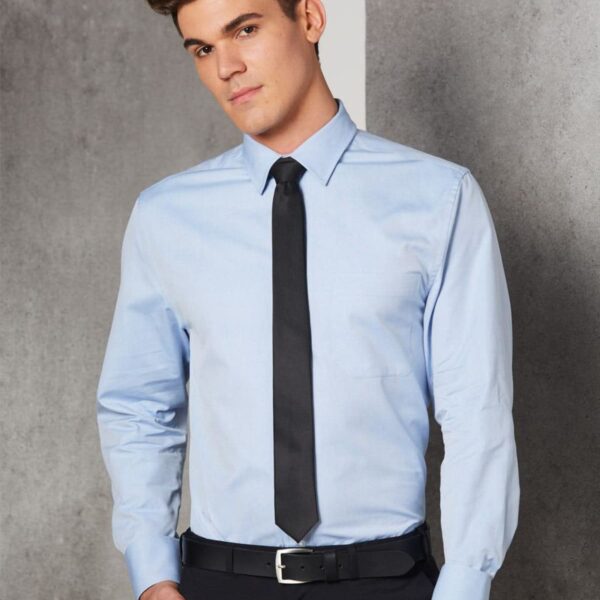 A man in a light blue dress shirt and black tie stands against a gray wall. His hands are in his pockets, and hes wearing black trousers with a matching belt.