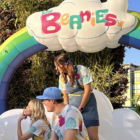 Three in colorful outfits under a Hexham Semi-circular Inflatables rainbow with The Beanies sign.