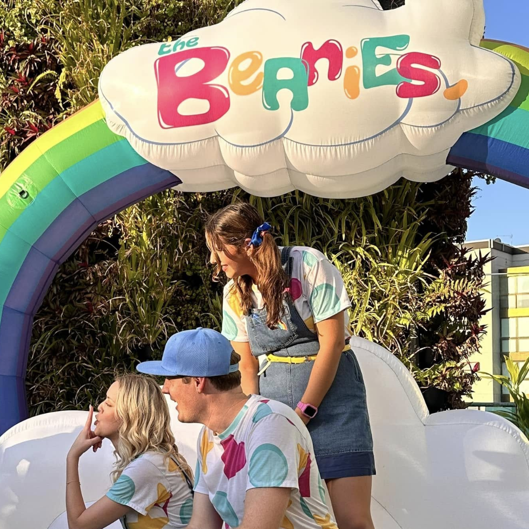 Three in colorful outfits under a Hexham Semi-circular Inflatables rainbow with The Beanies sign.
