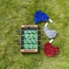 Mini foosball game on grass with red, blue, and silver pom-poms on each side.
