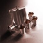 Metal flask with four small cups on a reflective surface.