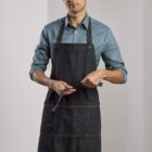 A person wearing a blue shirt and a dark apron stands indoors, holding the apron strap. The background is a softly lit interior with light-colored walls and wooden flooring. The individual looks towards the camera with a neutral expression.