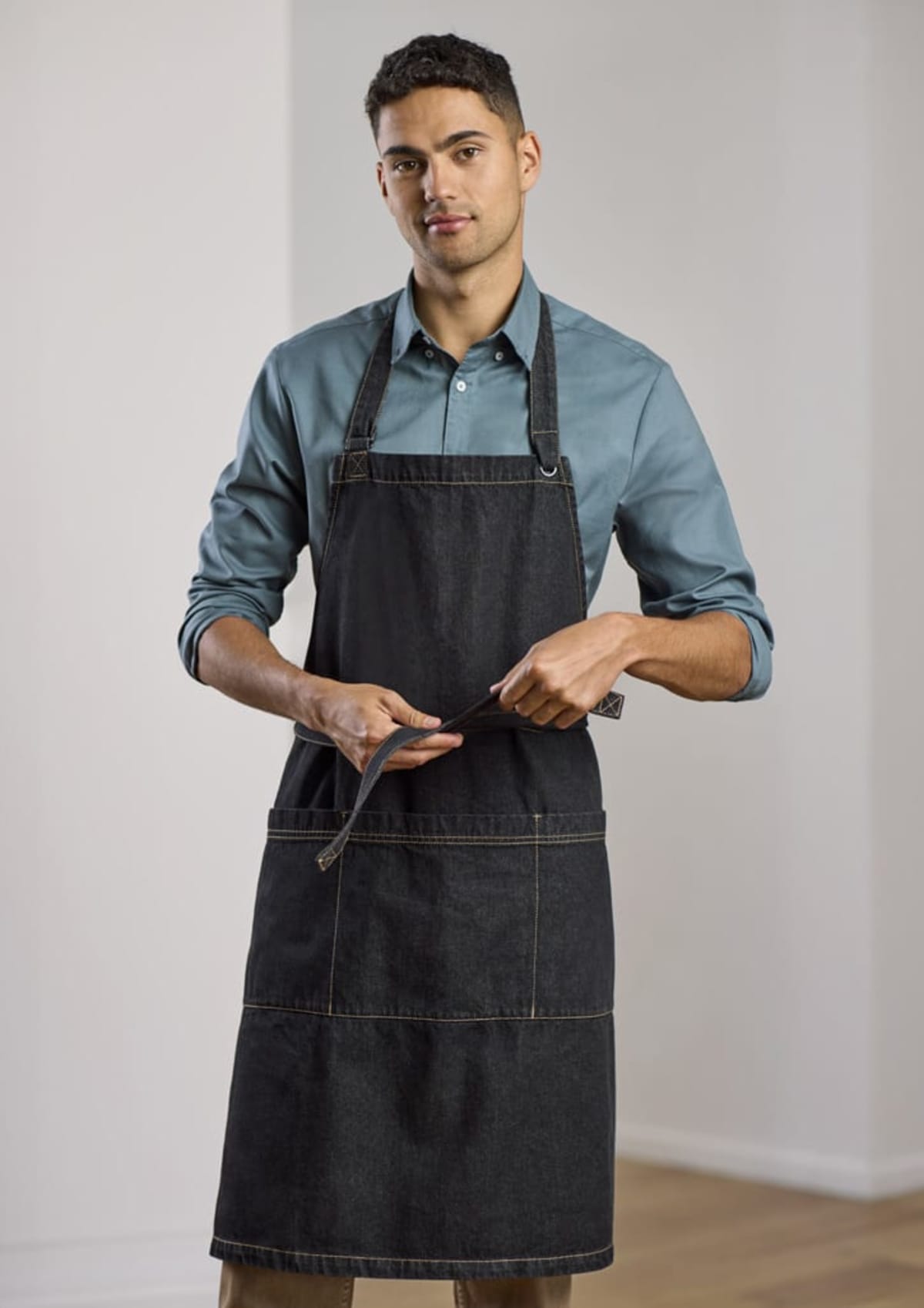 A person wearing a blue shirt and a dark apron stands indoors, holding the apron strap. The background is a softly lit interior with light-colored walls and wooden flooring. The individual looks towards the camera with a neutral expression.